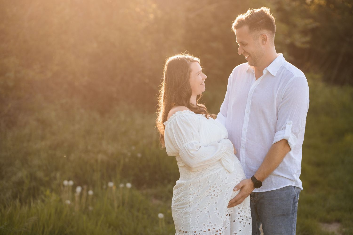 A couple embraces in a sunlit meadow during golden hour, wearing white and gray attire for their romantic photoshoot.