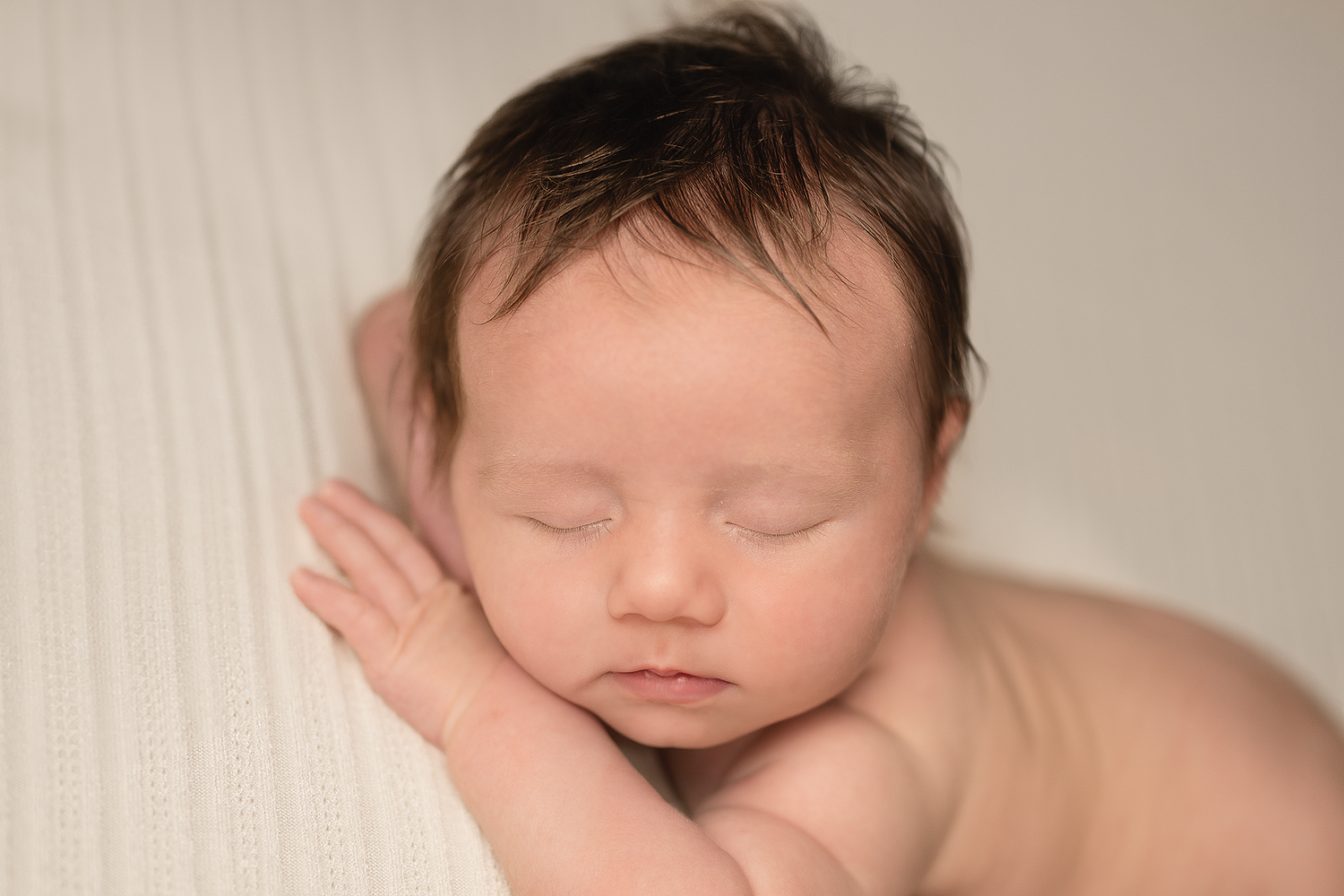 Peaceful newborn baby sleeping with dark hair, resting chin on tiny hands against a soft white background.