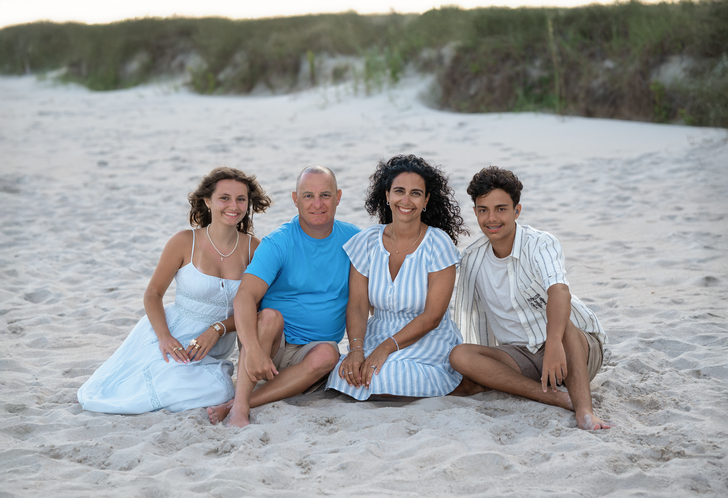 Family of four smiling together on a sandy beach, sitting casually in summer attire during golden hour.