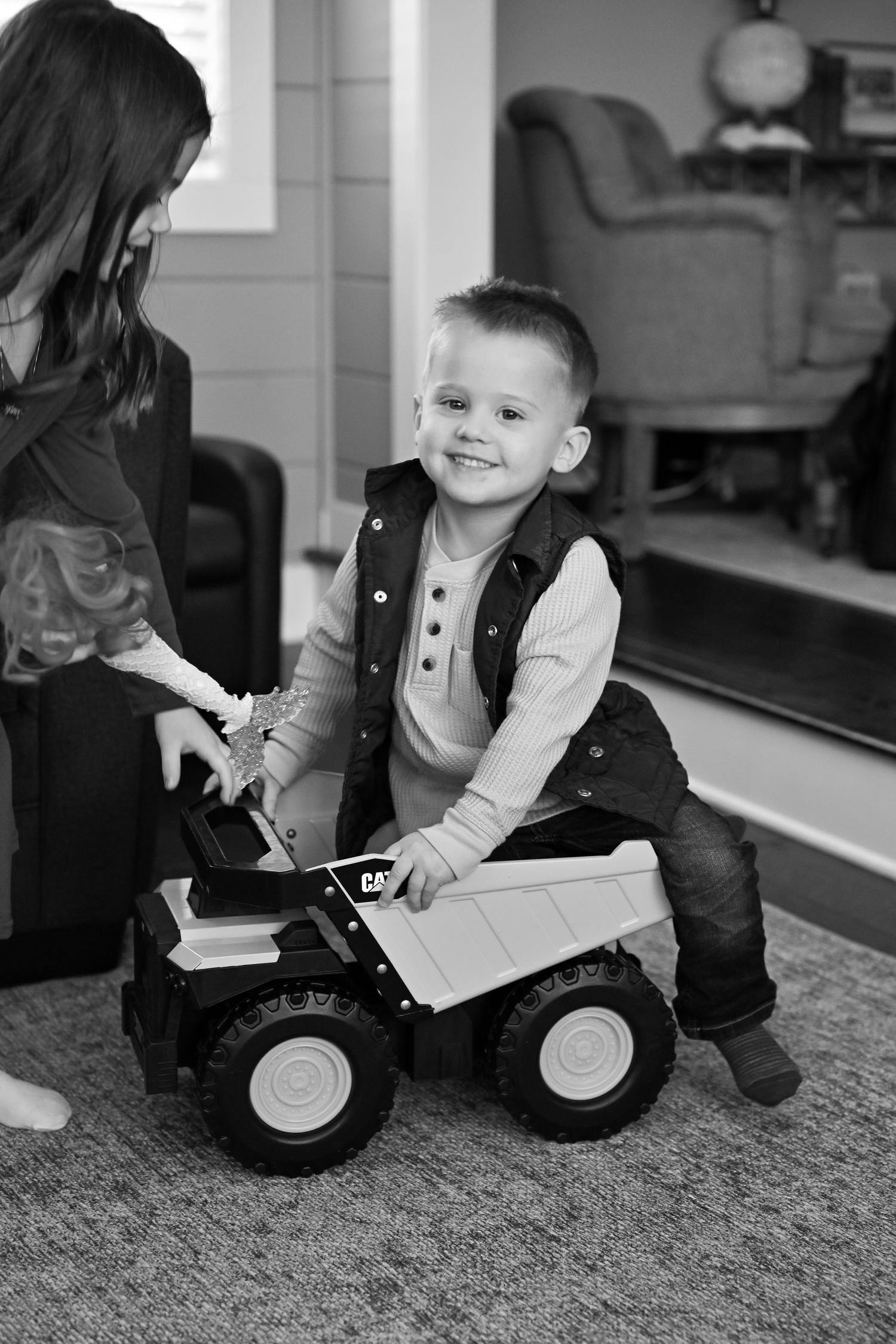 A smiling young boy plays with a large CAT toy dump truck on carpet while a girl watches nearby, in black and white.
