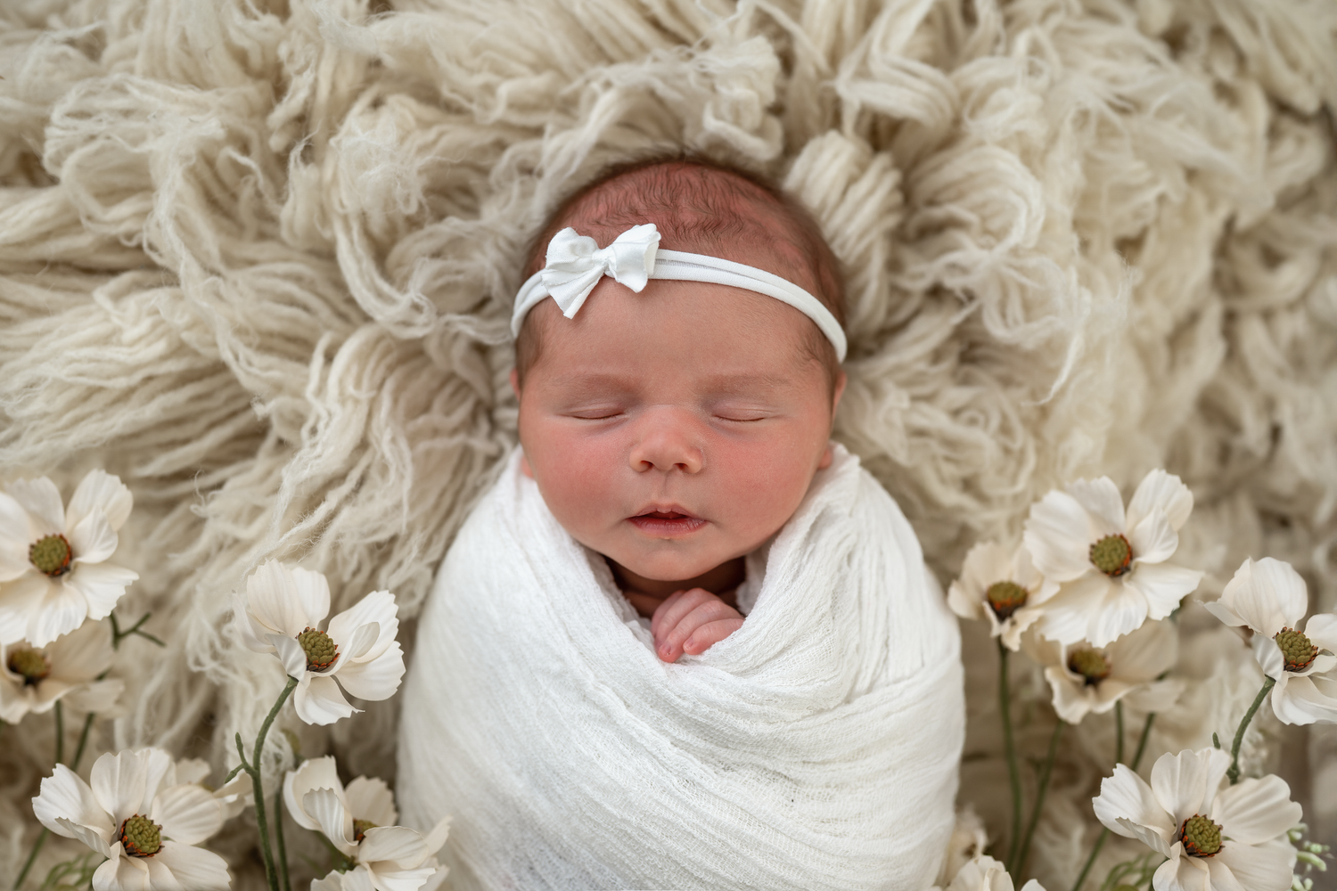Sleeping newborn baby swaddled in white wrap with bow headband, surrounded by white flowers on fluffy cream backdrop.
