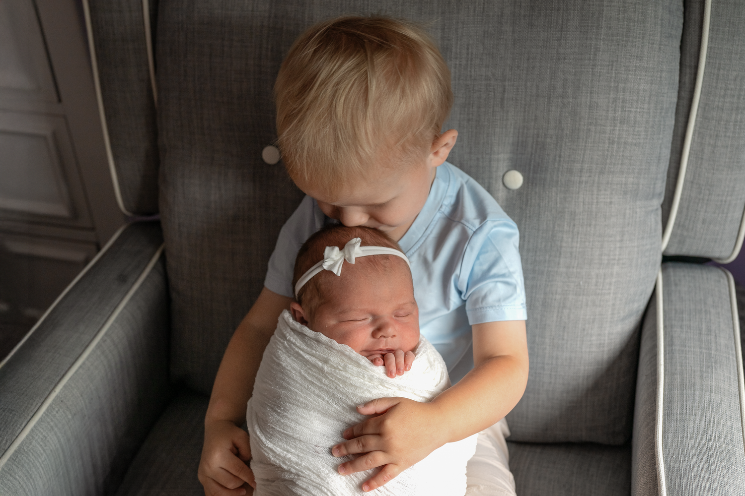 Toddler boy in blue shirt gently holds sleeping newborn baby swaddled in white on a gray chair.
