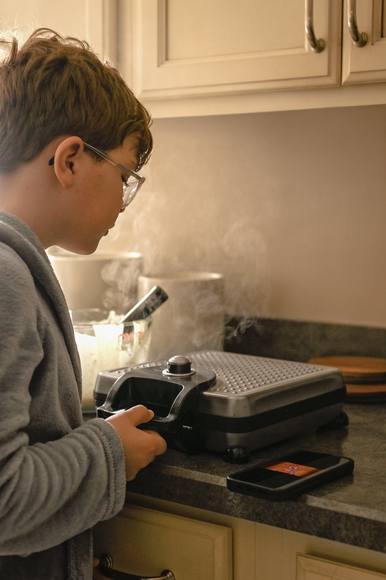 A person in a gray sweater uses a microscope on a black countertop in a kitchen setting.