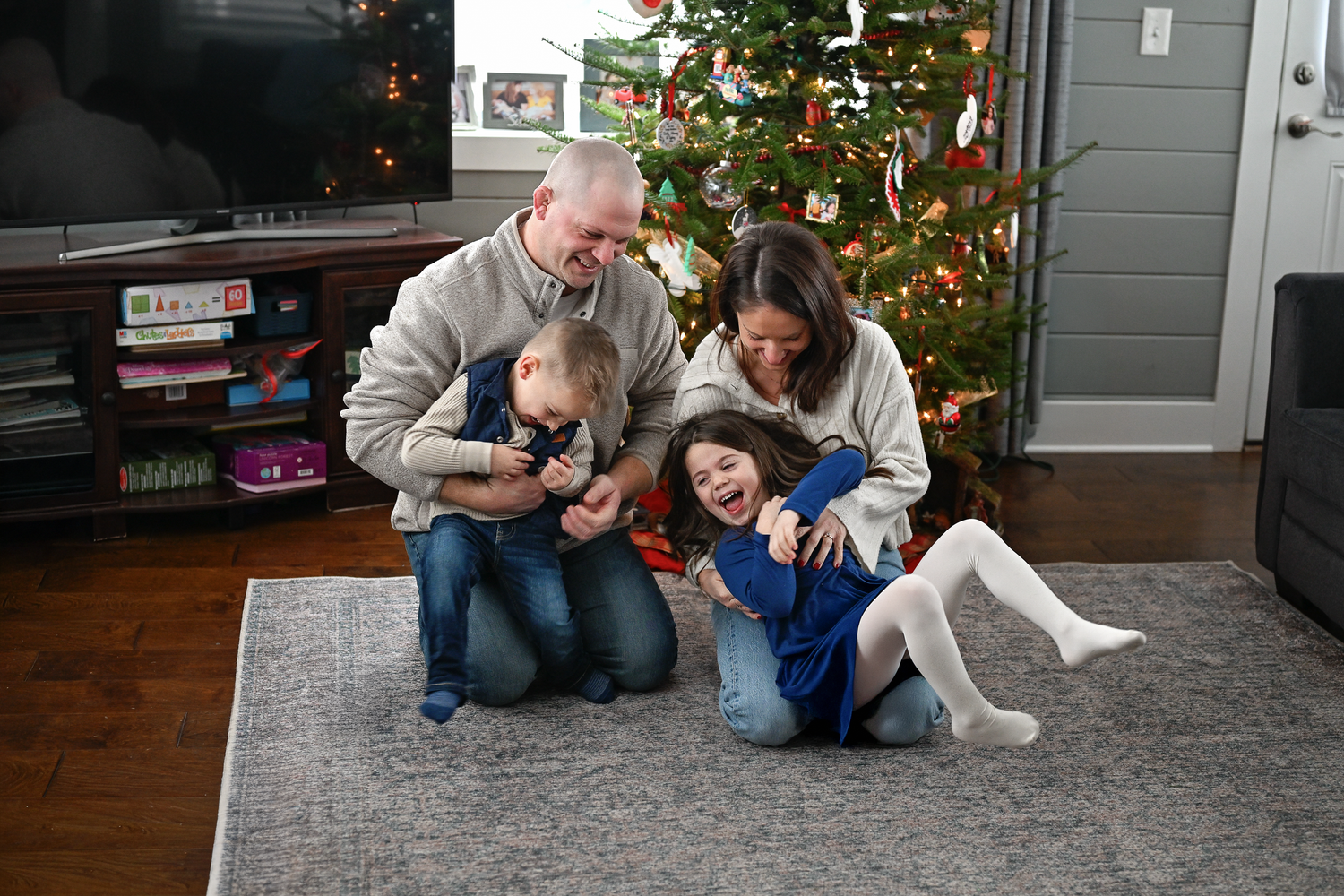 Family of four laughing together on living room floor in front of decorated Christmas tree during holiday season.