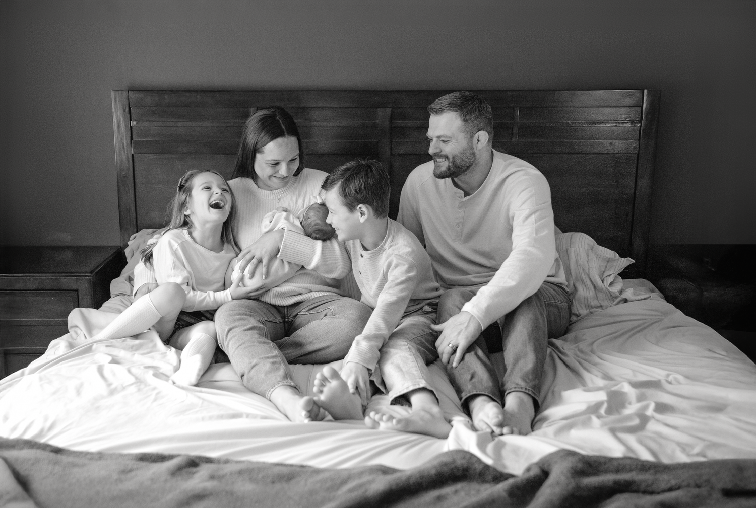 Black and white family photo of parents and three children laughing together on a bed.