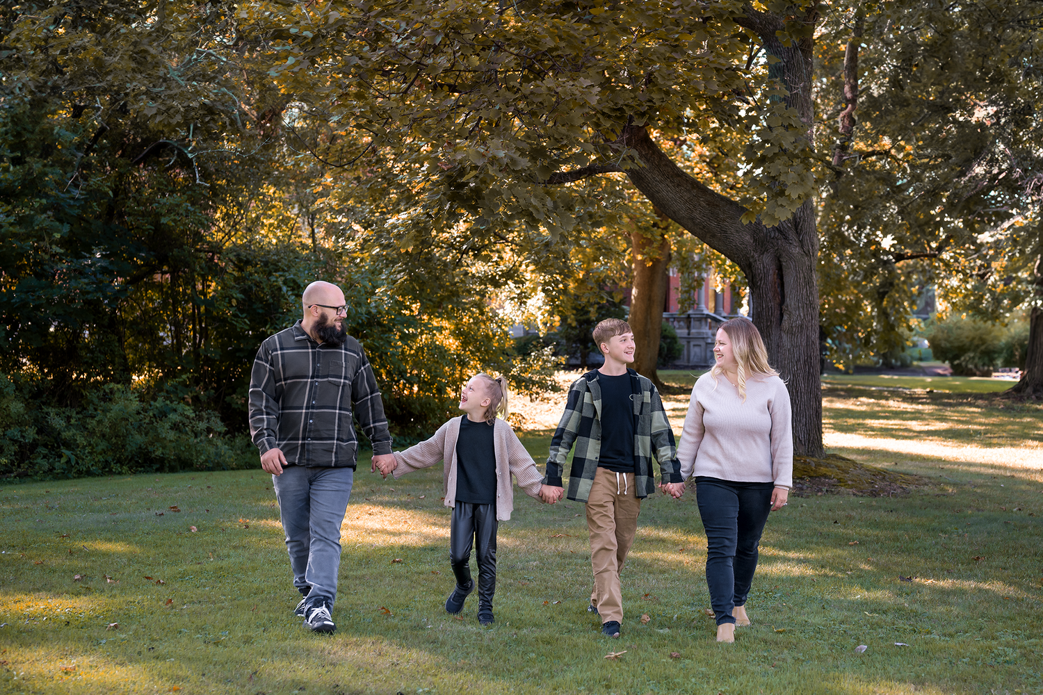 Family of four walking hand-in-hand through a lush green park surrounded by tall trees in autumn.