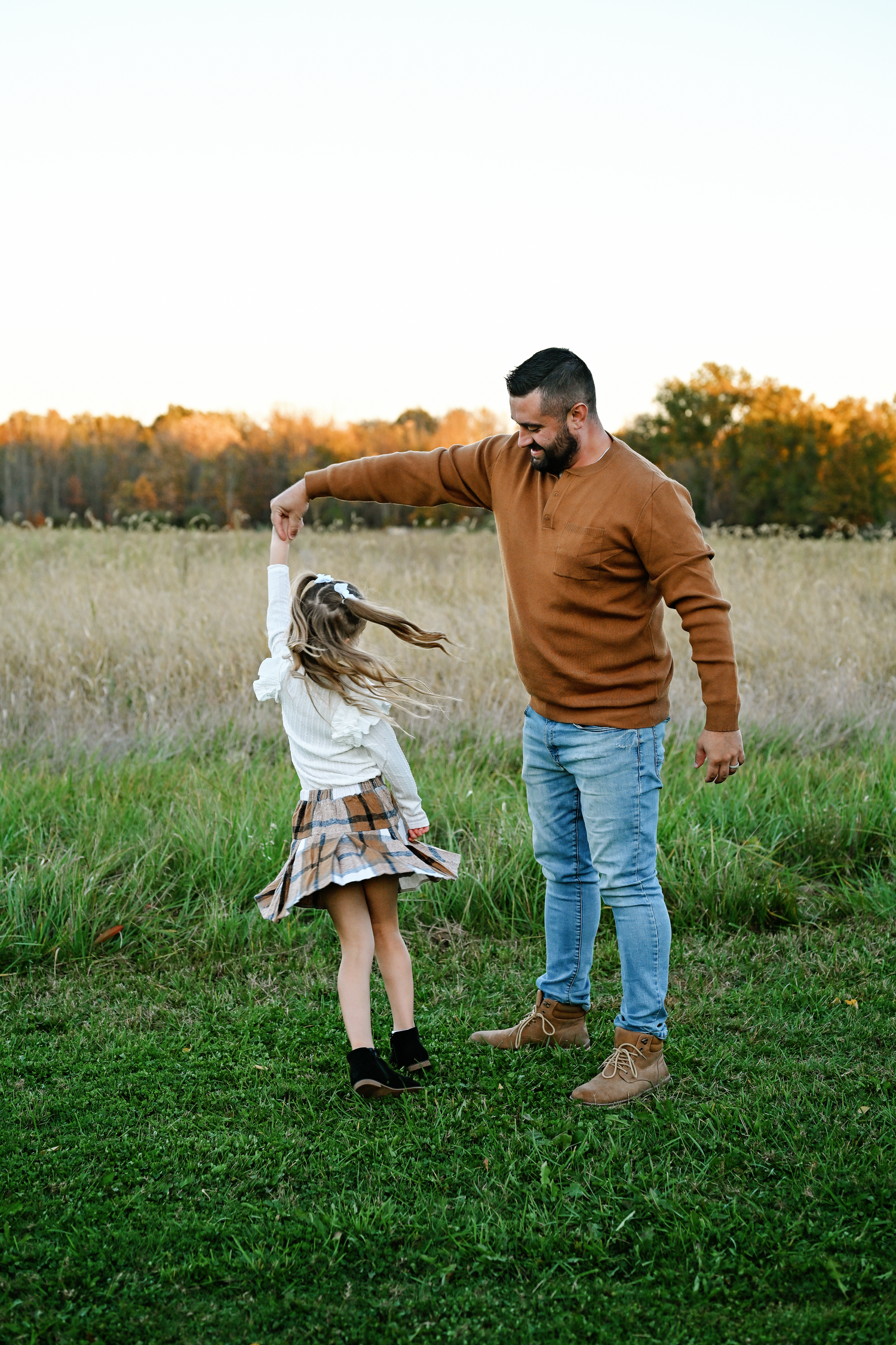 A father twirls his young daughter in a grassy field at sunset, both smiling in warm autumn outfits.