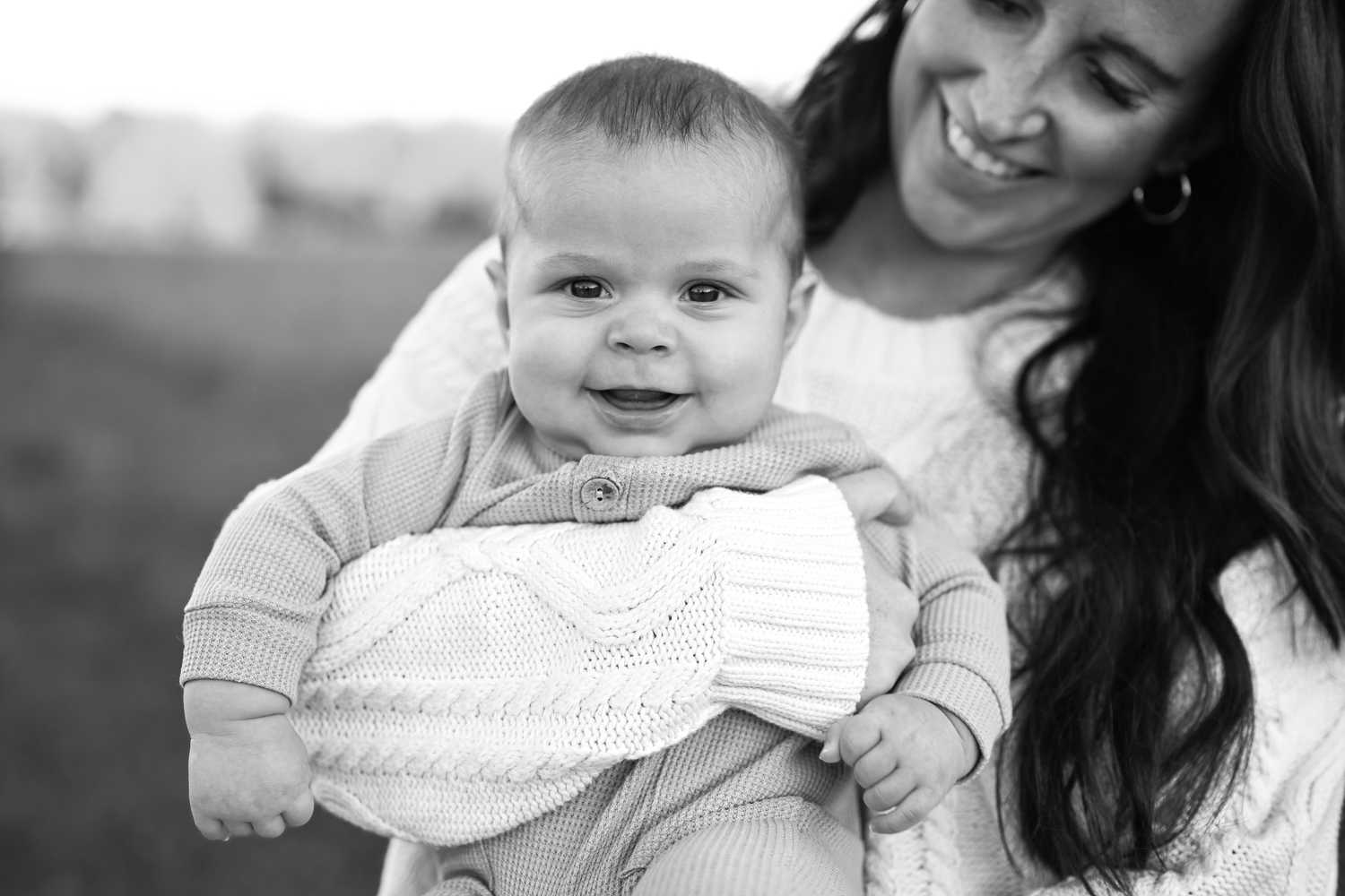 Black and white photo of a smiling baby held by a laughing woman outdoors.