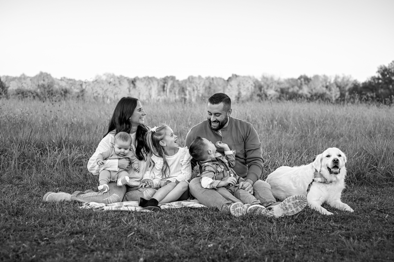 Black and white family photo of parents with two children and a golden retriever sitting in a grassy field.