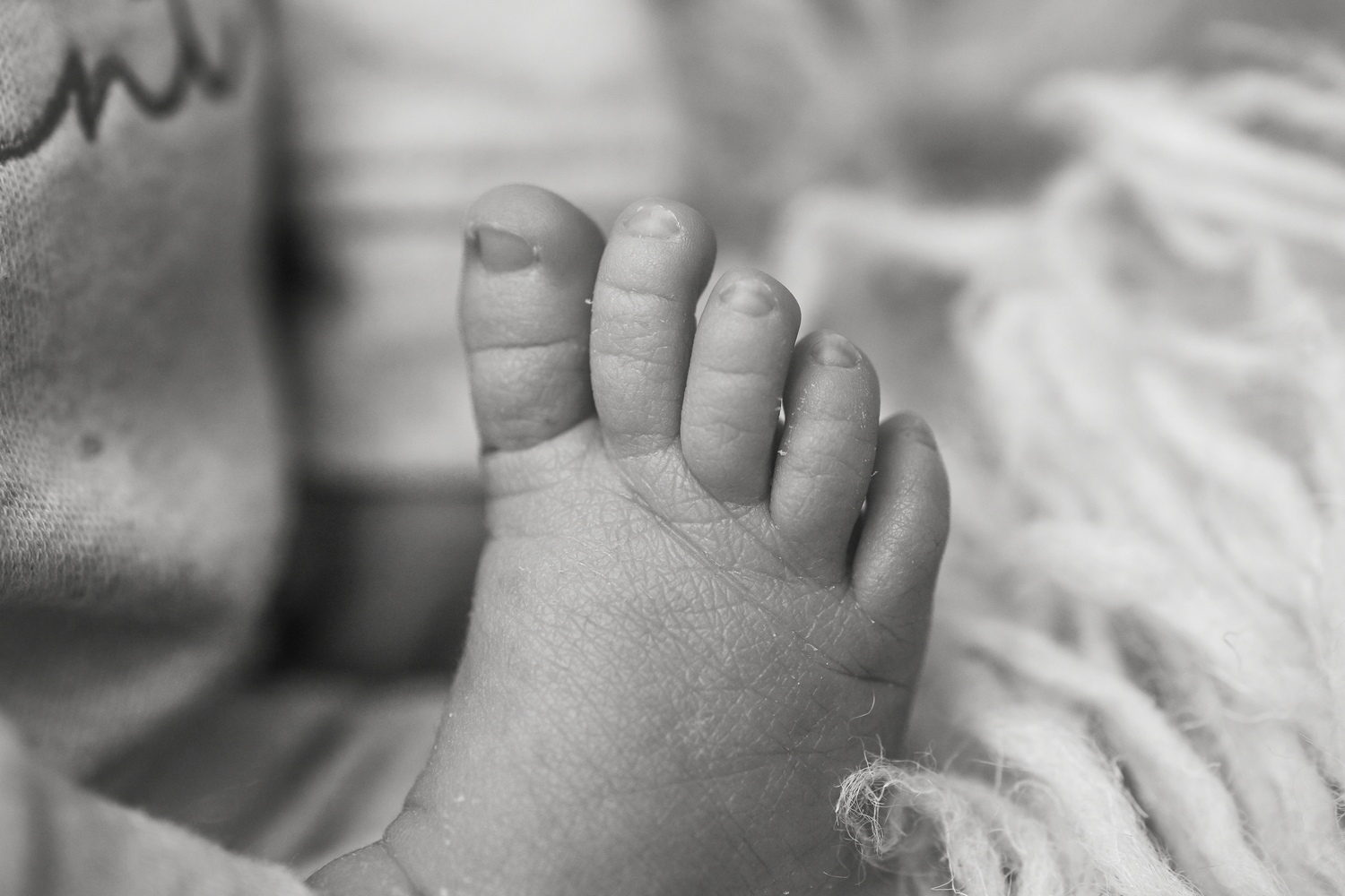 Close-up black and white photo of a newborn baby's tiny toes and foot resting on a soft fluffy white blanket.