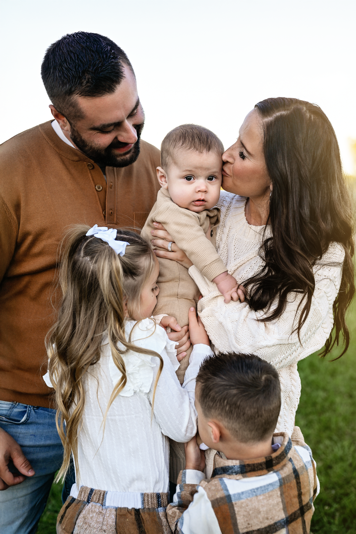Family of four with parents holding baby outdoors, mother kissing infant while children gather close in warm tones.