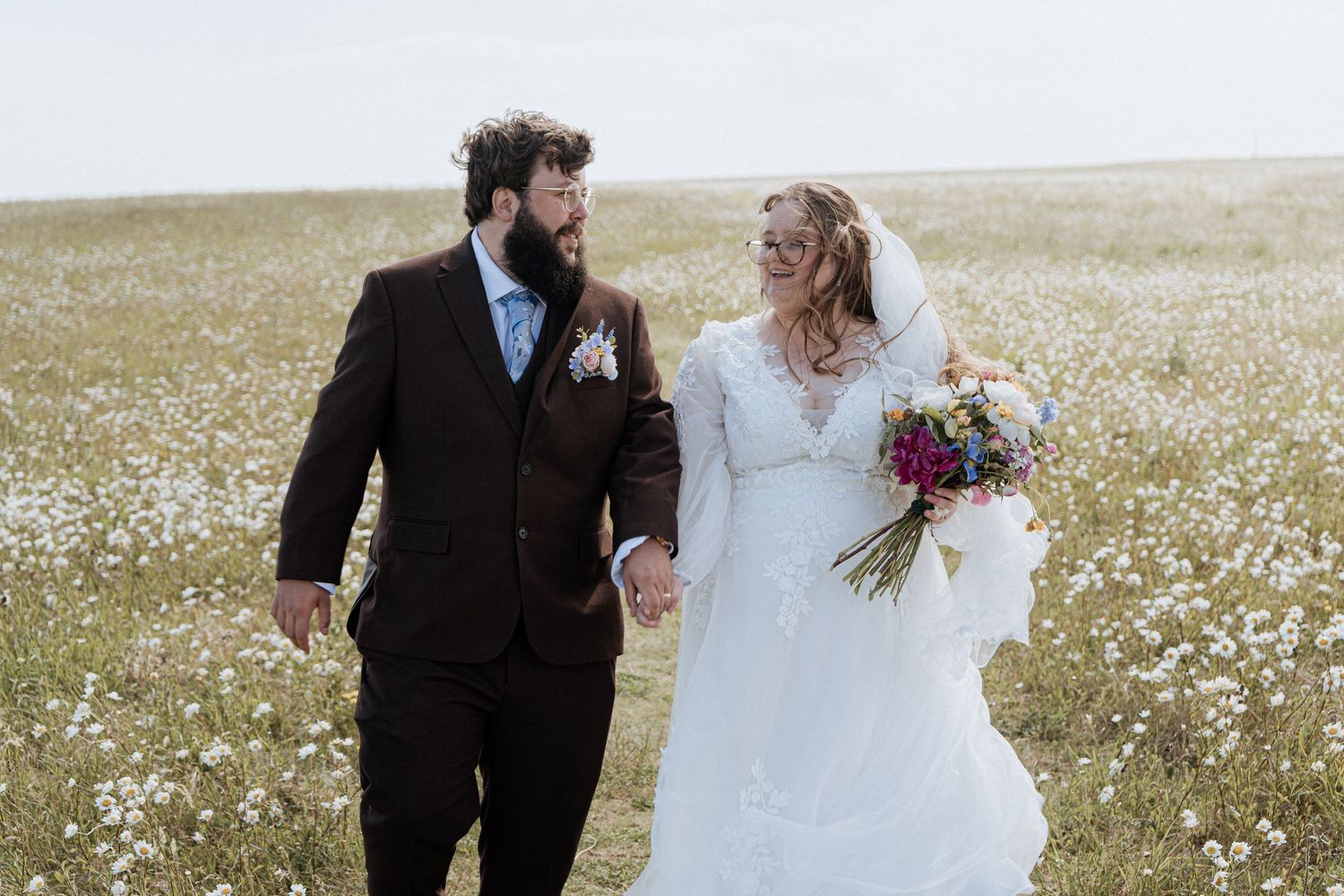 Newlywed couple holding hands walking through a field of white wildflowers on their wedding day.