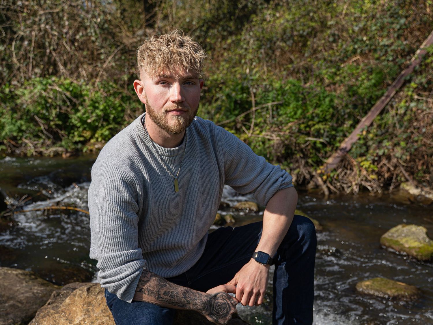 Bearded man in a grey sweatshirt sitting on rocks beside a woodland stream in a natural outdoor setting.