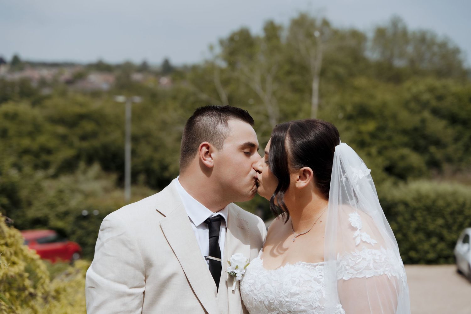 Bride and groom sharing a kiss outdoors with green trees and urban landscape in background.