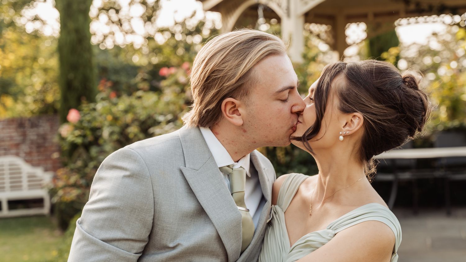 A couple shares a romantic kiss at an outdoor wedding, groom in gray suit, bride in elegant updo hairstyle.