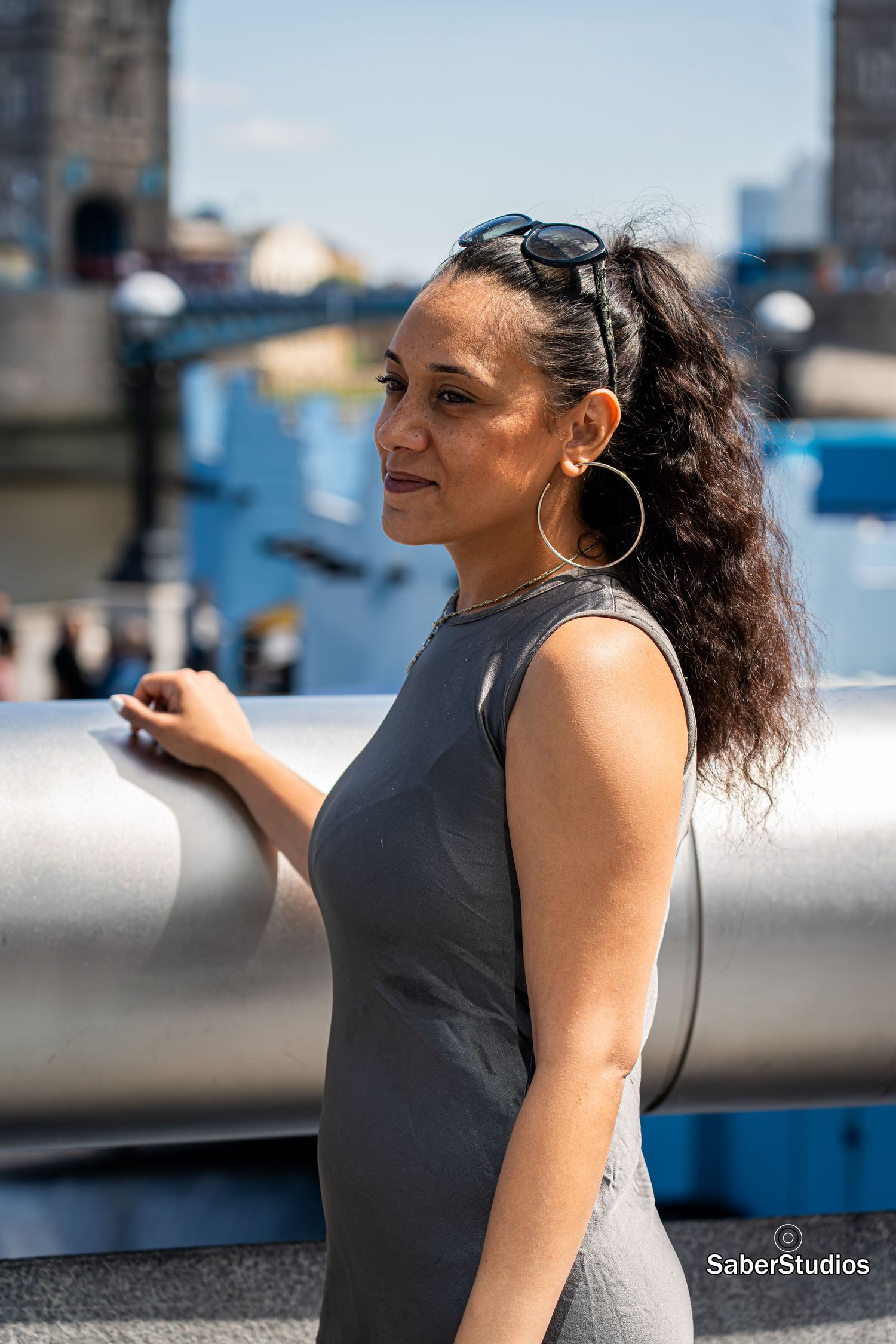 Woman in a sleeveless grey dress with curly hair and hoop earrings posing near a railing with Tower Bridge behind.