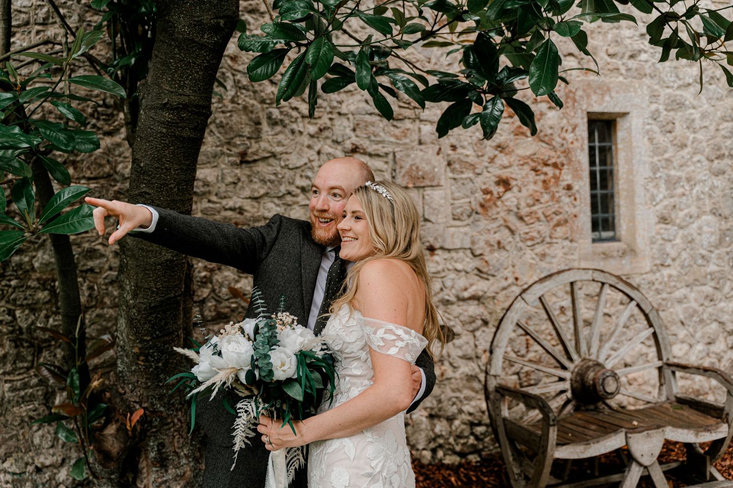 Bride and groom pose playfully under tree beside stone wall and rustic wooden wagon wheel.
