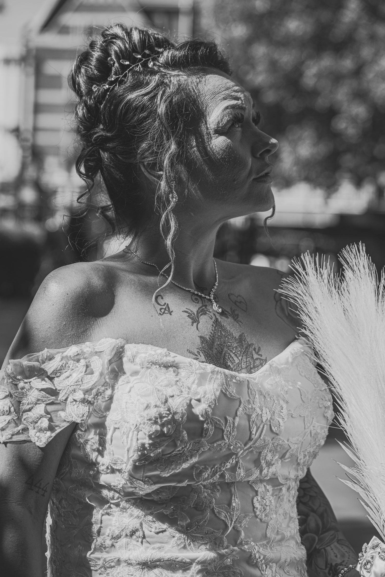 Black and white portrait of a bride in an off-shoulder lace gown with updo hairstyle and tattoo, holding feather bouquet.