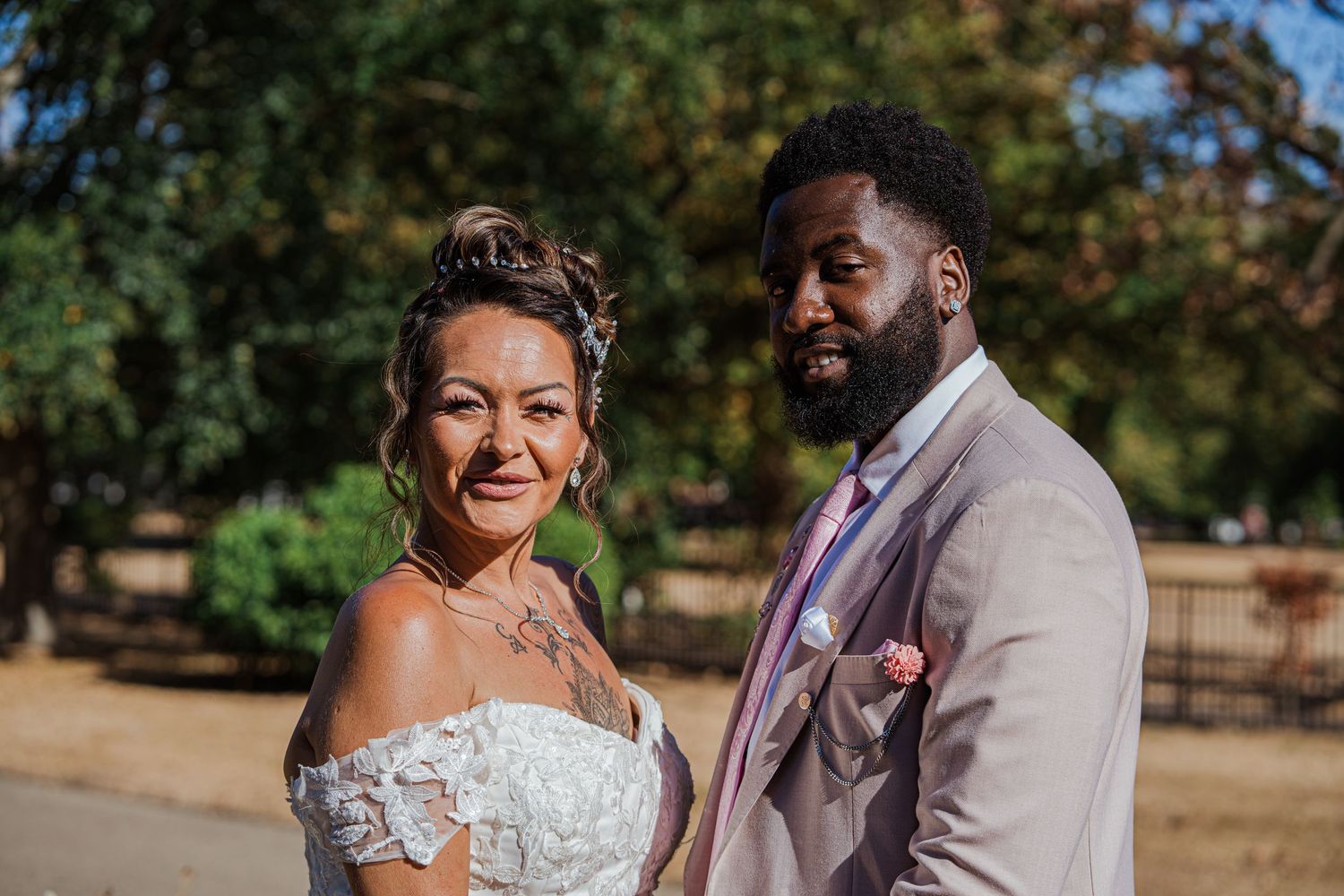 A bride and groom pose outdoors in a park, she in a lace gown and he in a gray suit, on their wedding day.