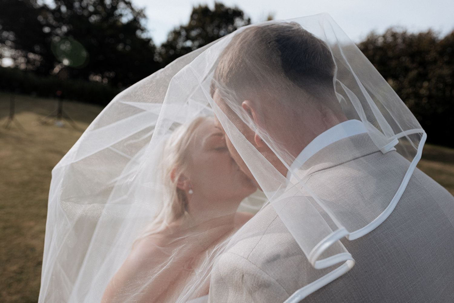Bride and groom share romantic kiss under sheer wedding veil in soft natural light.
