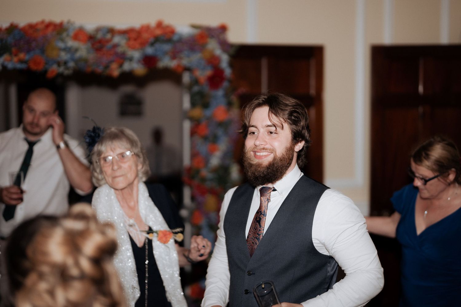 Bearded groom in gray vest smiles at wedding reception with elderly guest and colorful floral backdrop.