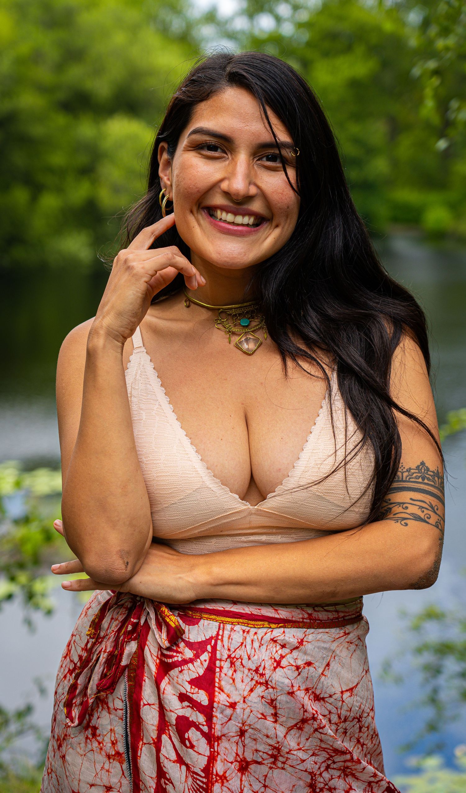Smiling woman with long black hair wearing a beige bralette top and floral skirt, posing outdoors near a lake.