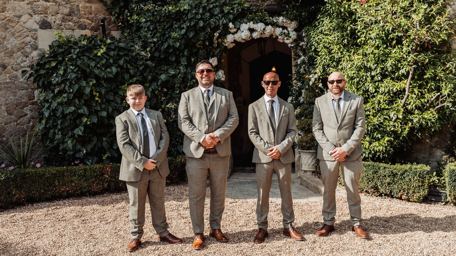 Four groomsmen in grey suits with sunglasses standing outside floral-adorned stone doorway.