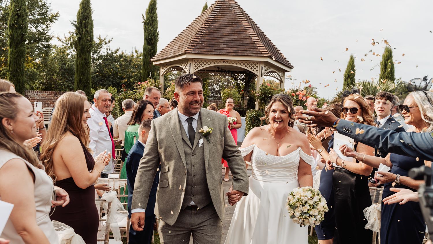 Happy couple exits wedding ceremony through confetti toss near elegant garden gazebo.
