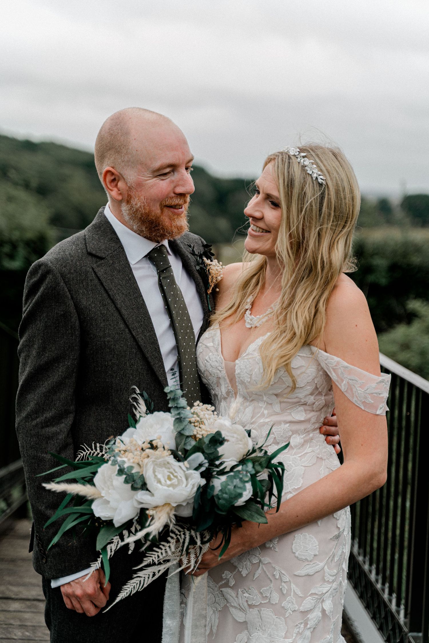 A smiling bride and groom share a romantic moment on a bridge, she holding a white floral bouquet on their wedding day.
