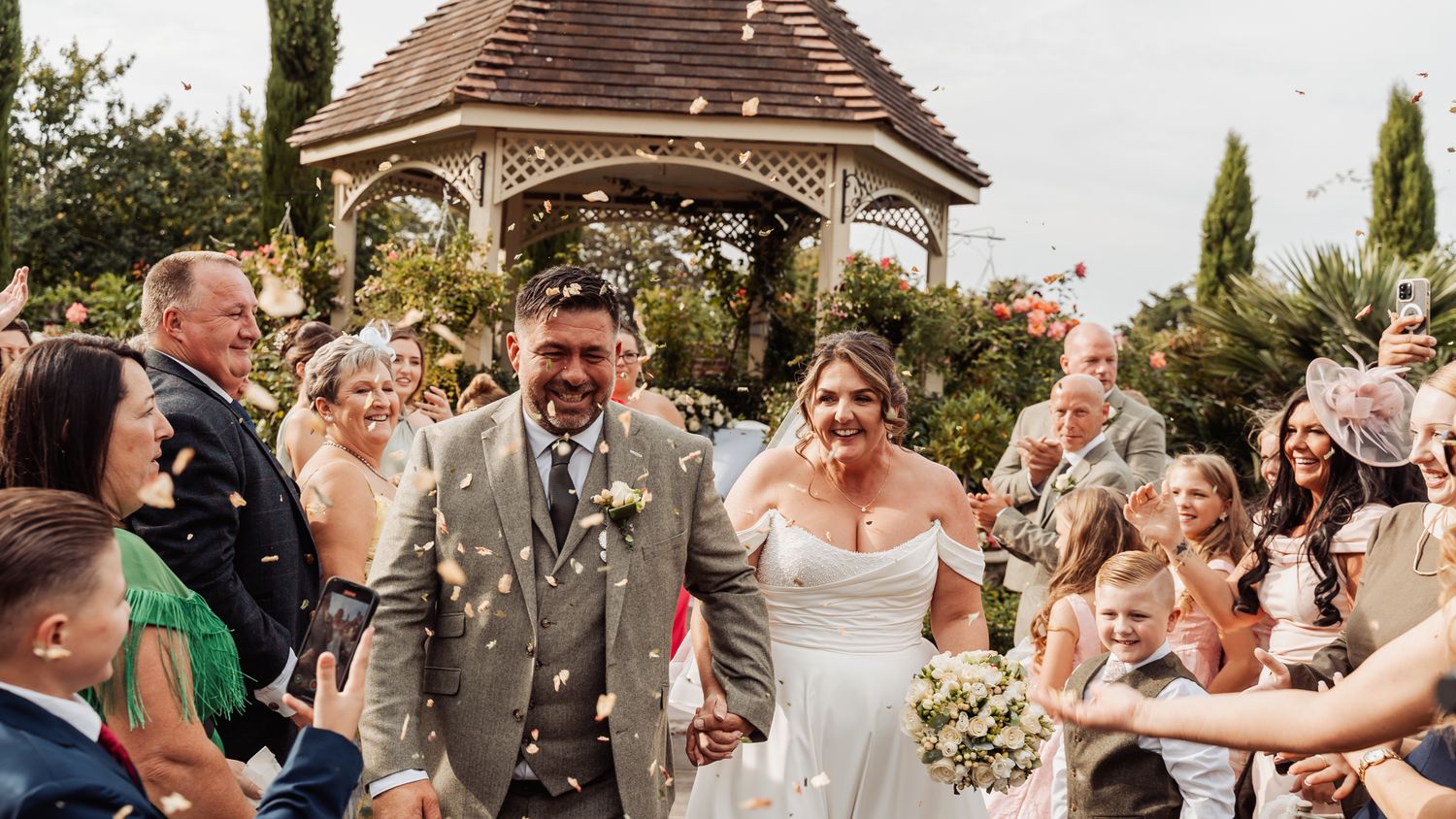 Bride and groom walk through confetti shower with guests cheering outside garden gazebo.