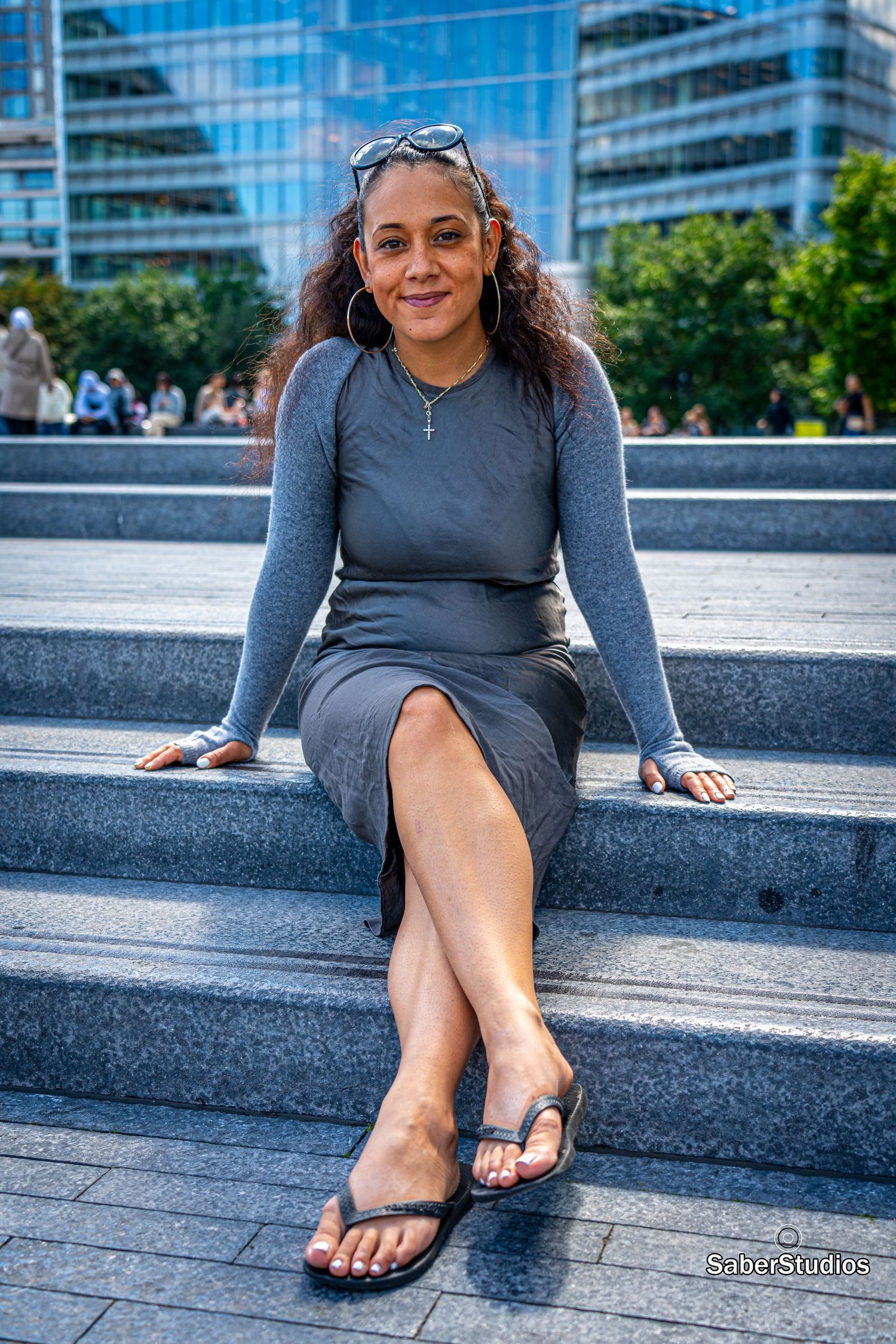 Curly-haired woman in a grey long-sleeve dress sitting on stone steps in an urban setting with glass buildings.