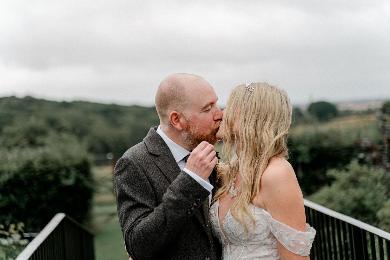 Bald groom in grey suit kissing blonde bride on outdoor balcony with scenic countryside view.