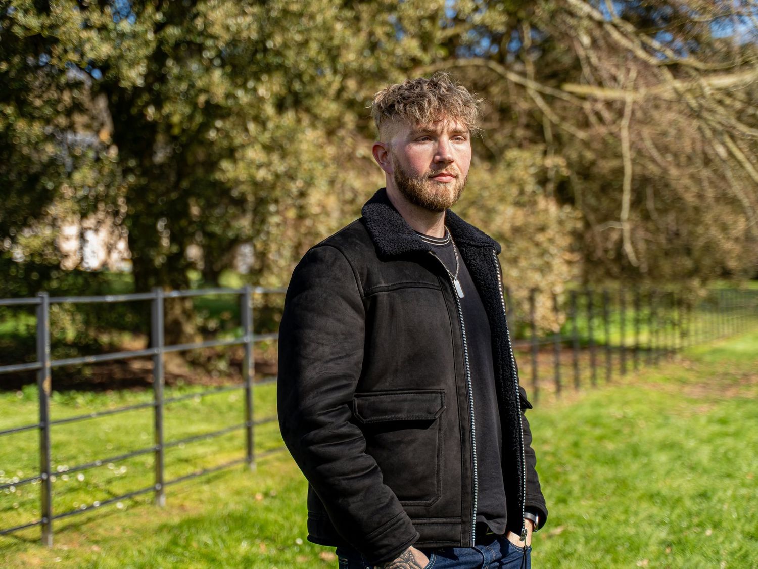 Bearded man in a black jacket standing in a green field with trees and a fence visible in the background.