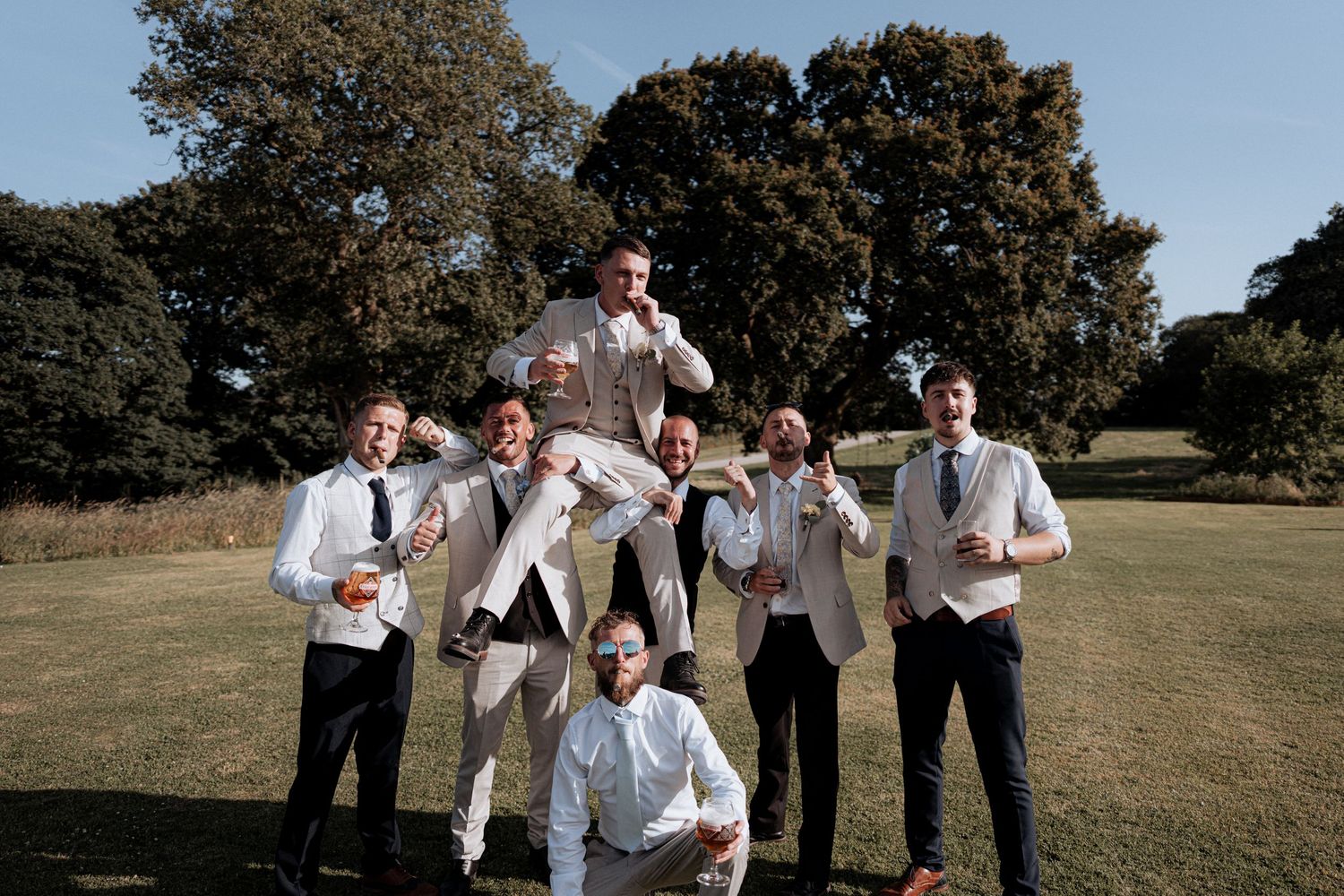 Groom lifted by groomsmen in beige suits on sunny green lawn with trees in background.