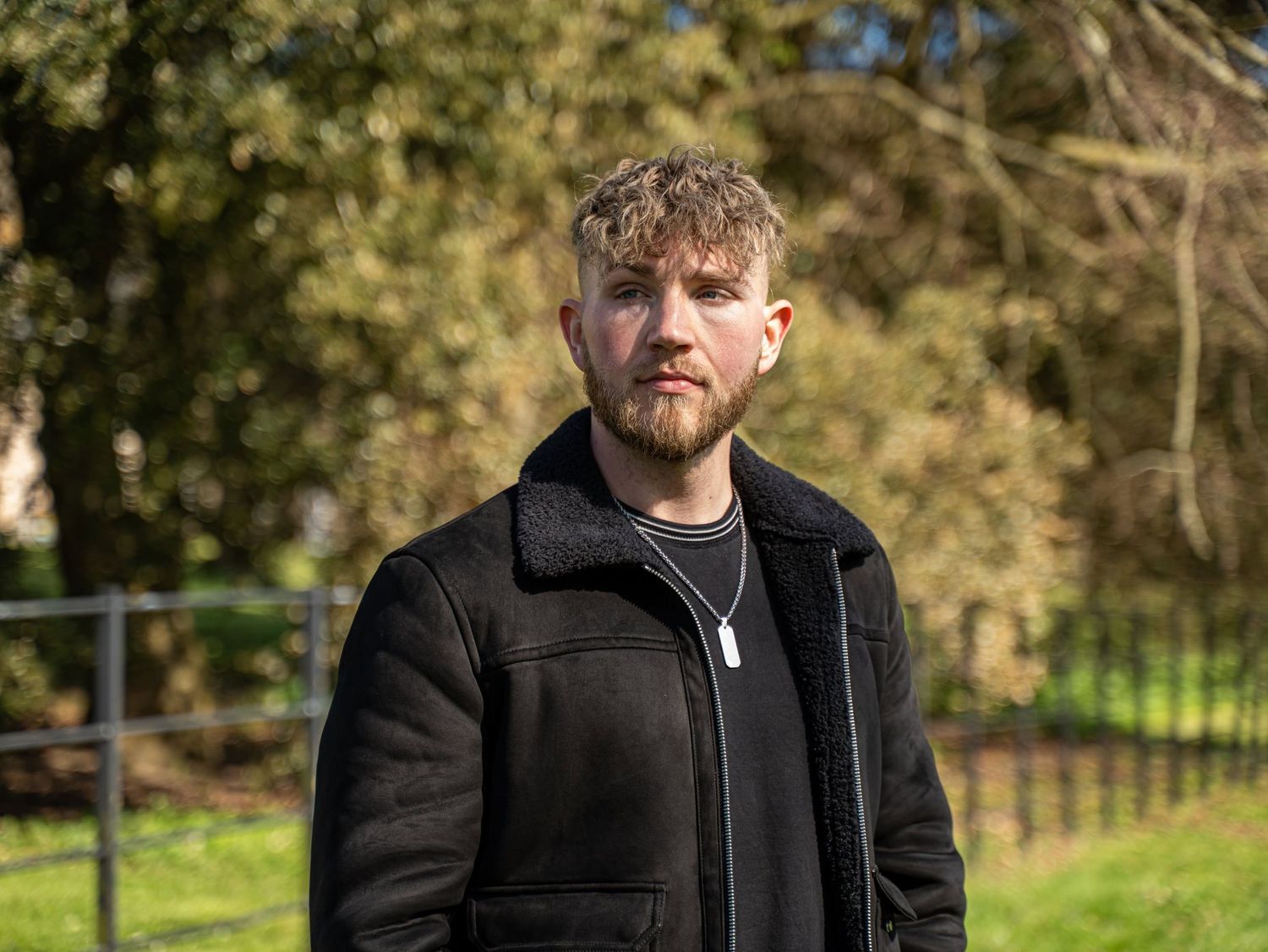 Young bearded man in a black shearling jacket standing outdoors in a park with trees in the background.