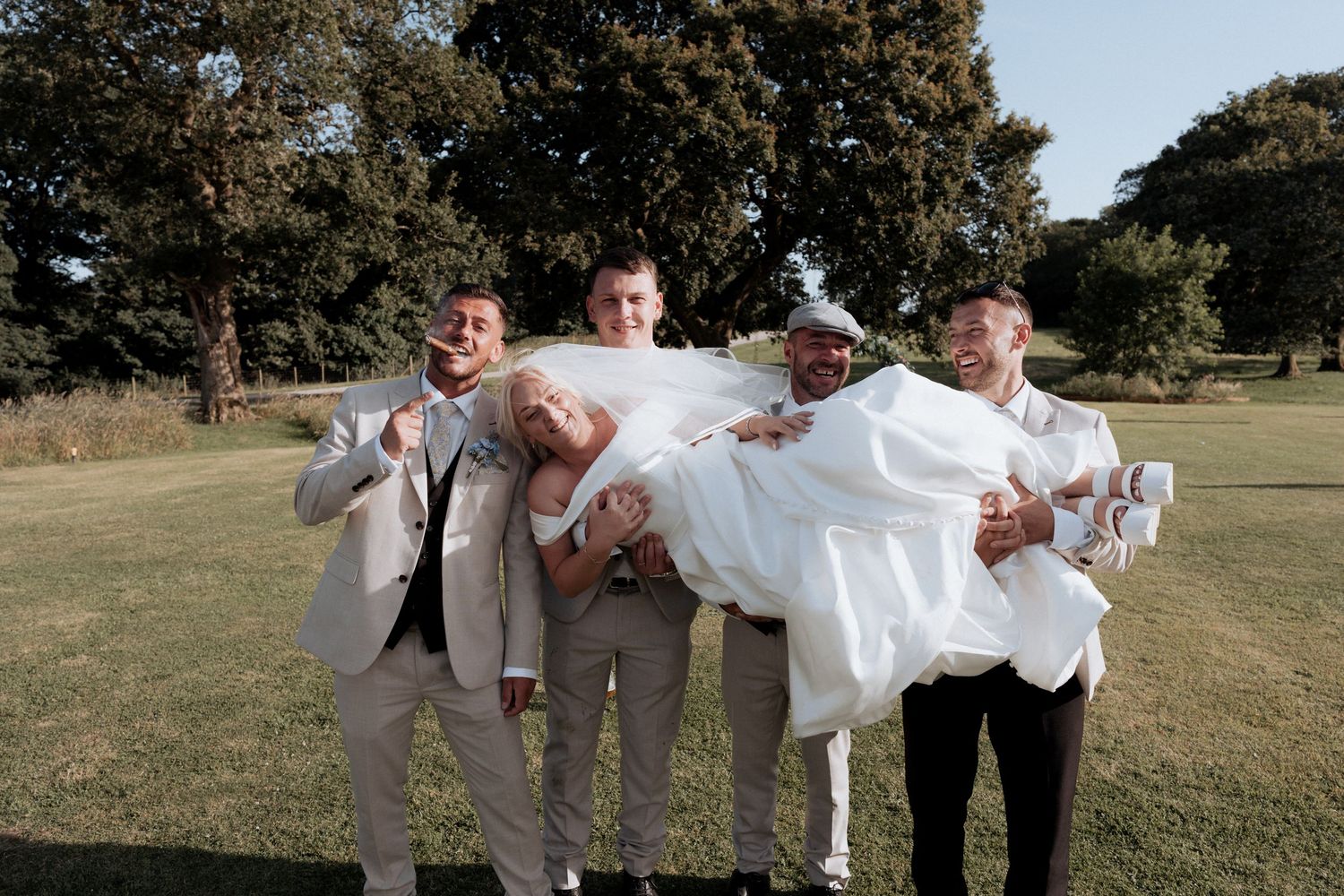 Groomsmen carry laughing bride across sunny green lawn in playful wedding party photo.