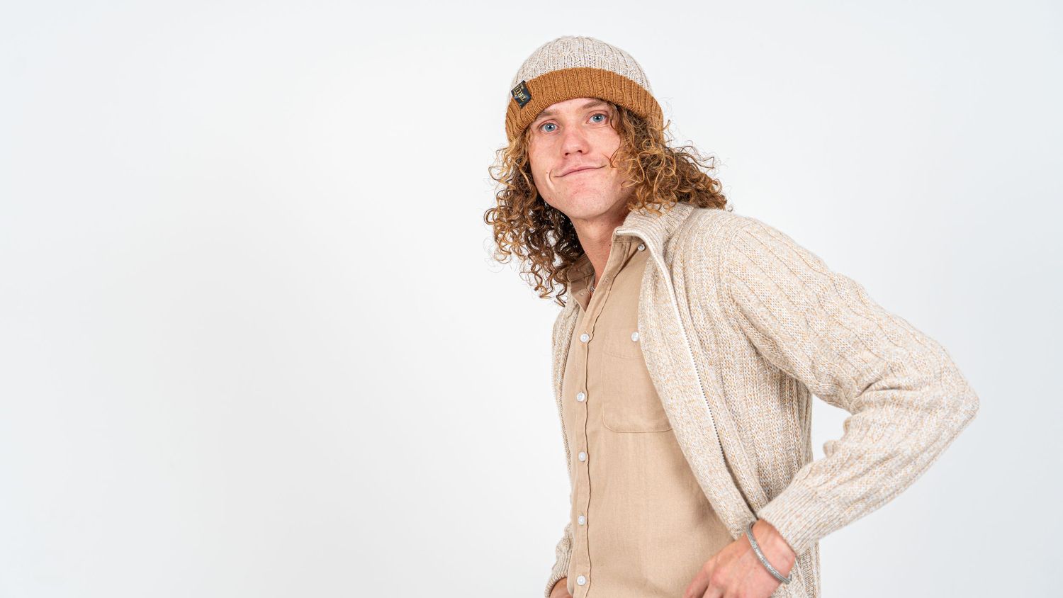 Young man with curly hair wearing beige knit outfit and brown beanie posing on white background.