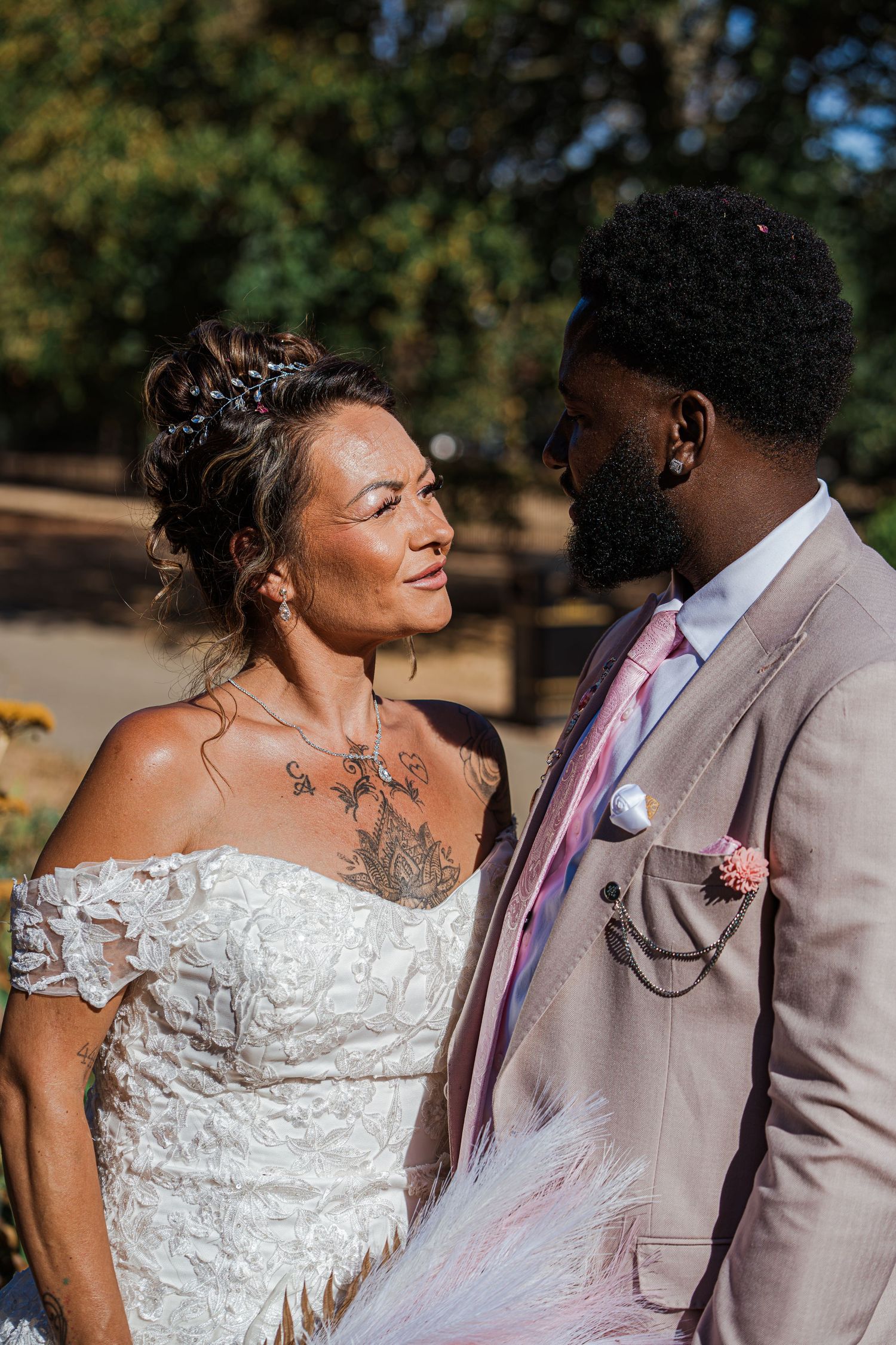 Interracial couple in wedding attire share a loving gaze outdoors, bride in lace gown, groom in pink suit.