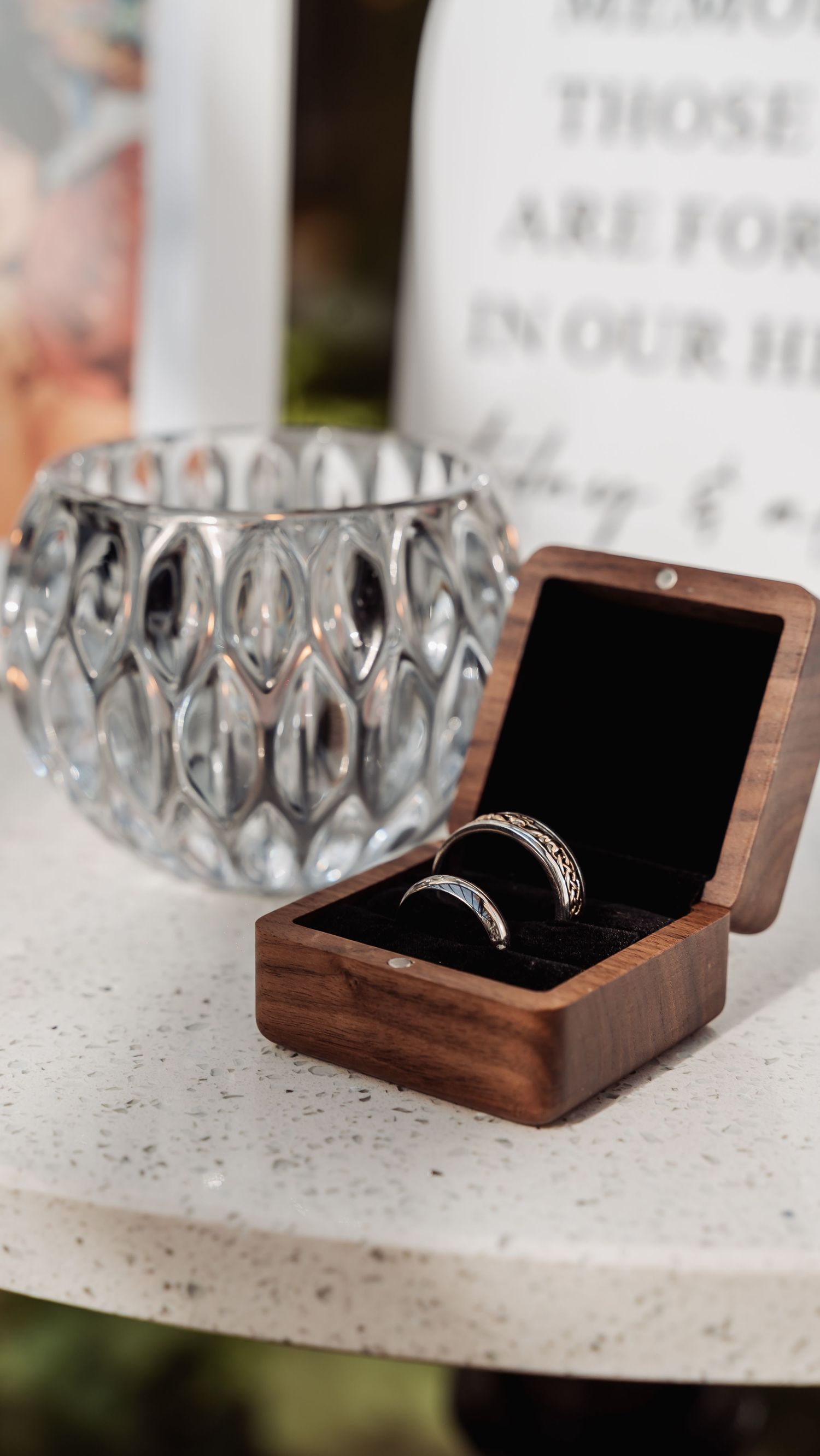Two silver wedding bands displayed in wooden ring box beside crystal votive holder.