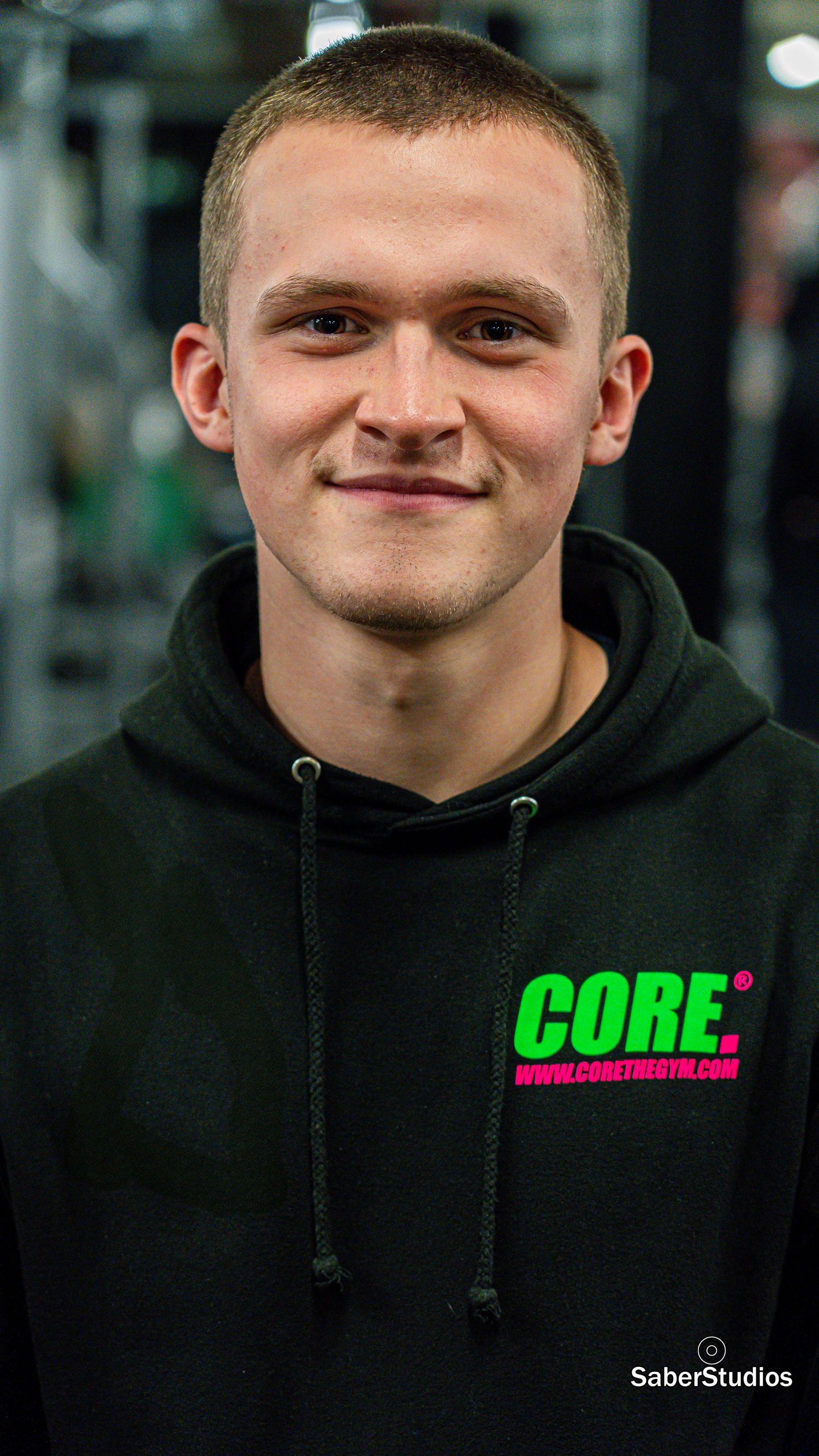 Young man smiling in a black Core gym hoodie with neon green and pink logo inside a fitness center.