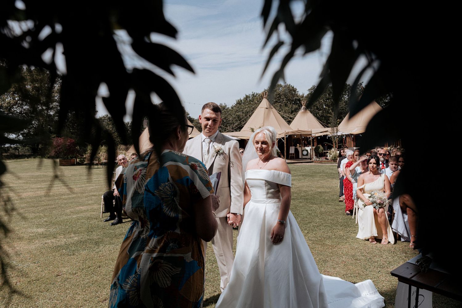 Newlyweds greet guests outside tipi wedding venue on sunny day with wedding party nearby.