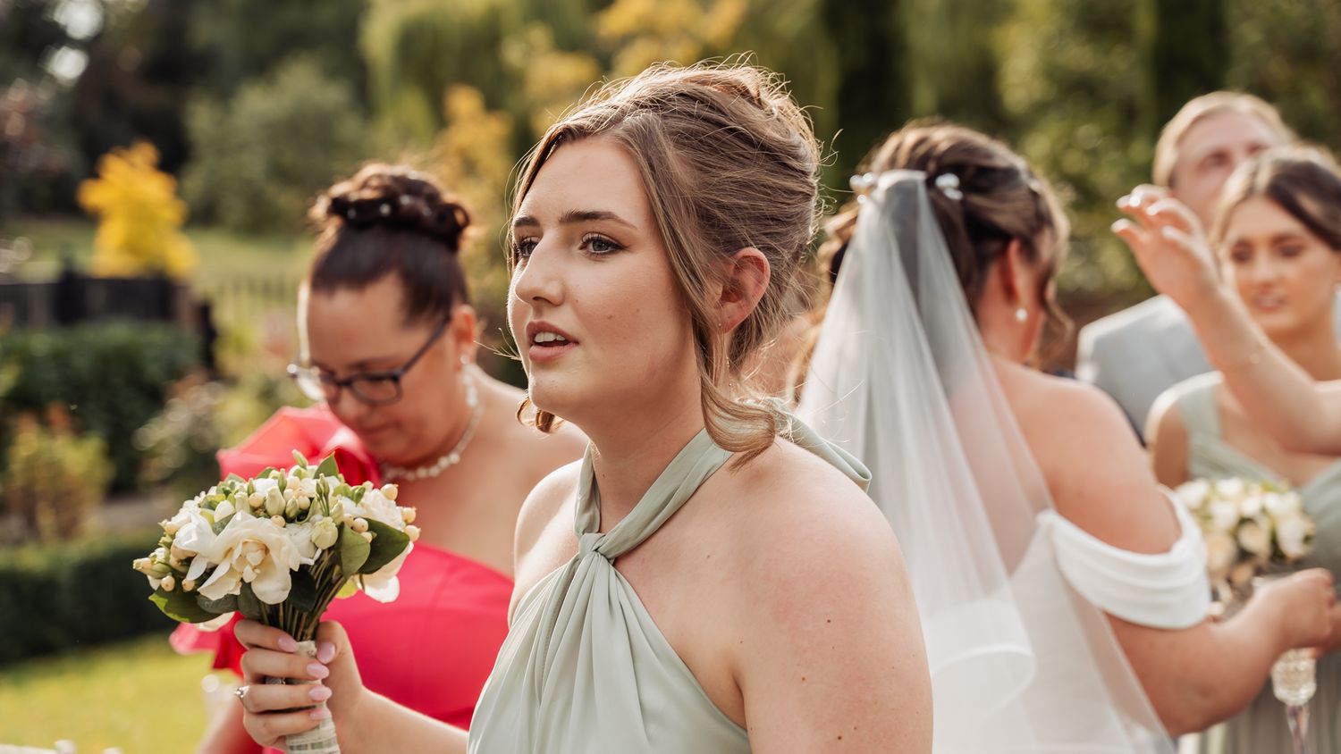 Bridesmaid in sage green dress holds white bouquet during outdoor wedding ceremony.