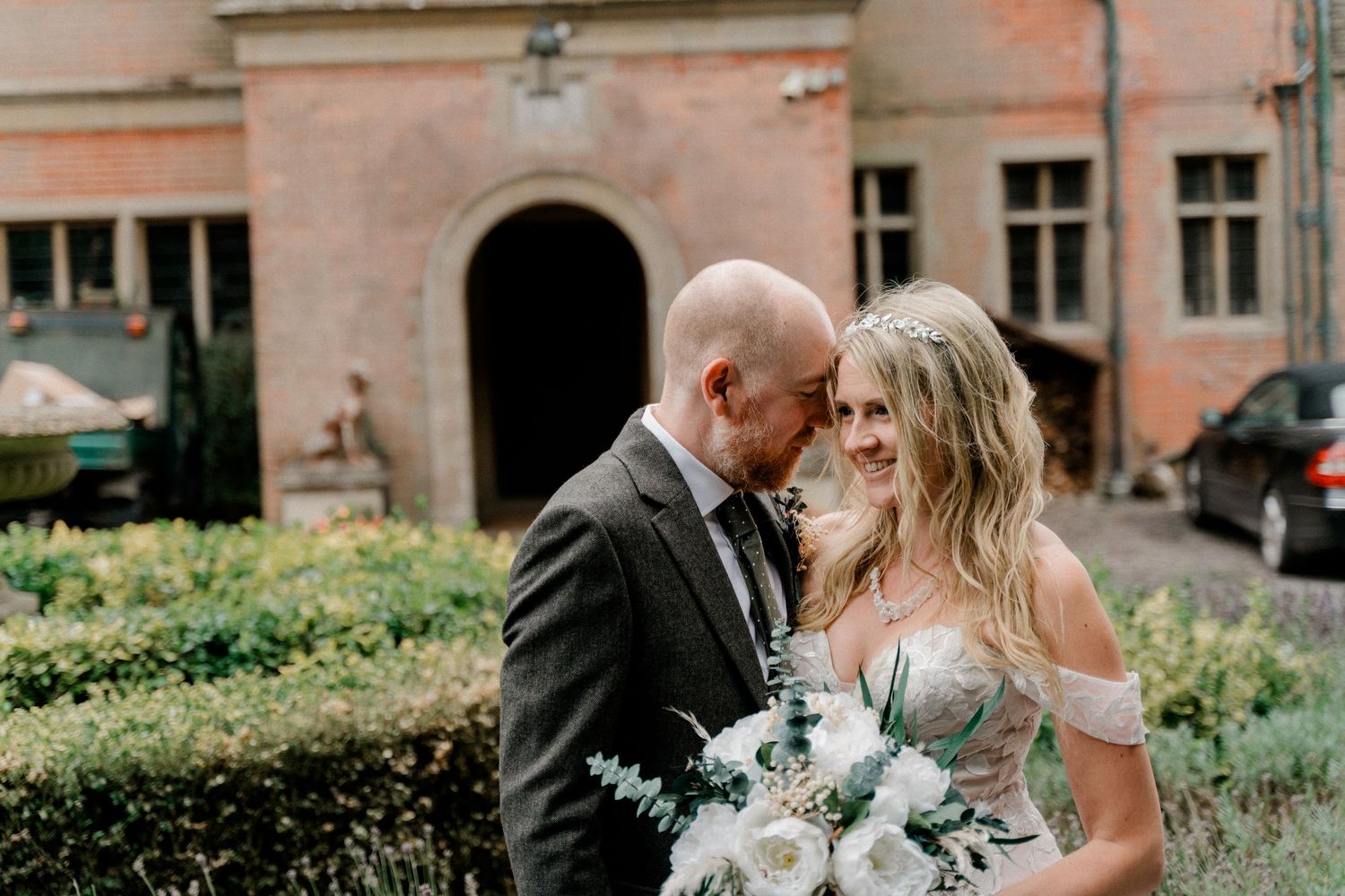 Bride and groom share intimate moment outside historic stone building with white floral bouquet.