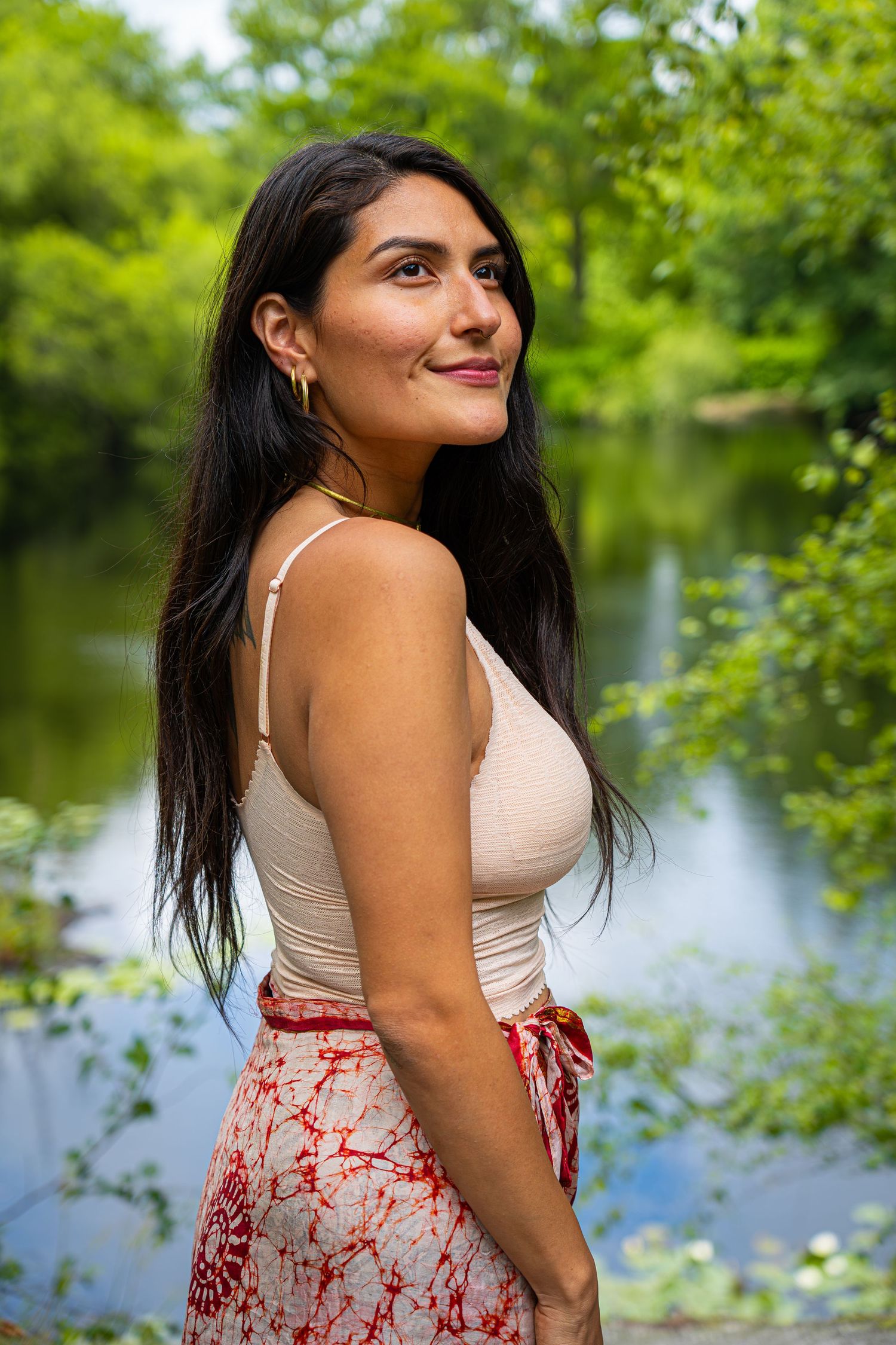 Brunette woman in a beige cami top and red tie-dye skirt posing by a serene lake surrounded by lush trees.