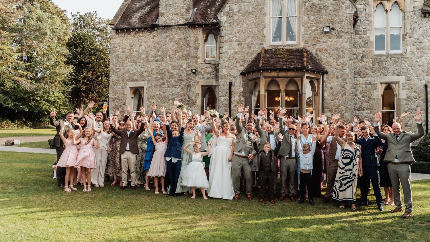 Large wedding party group photo with hands raised outside historic stone manor house.