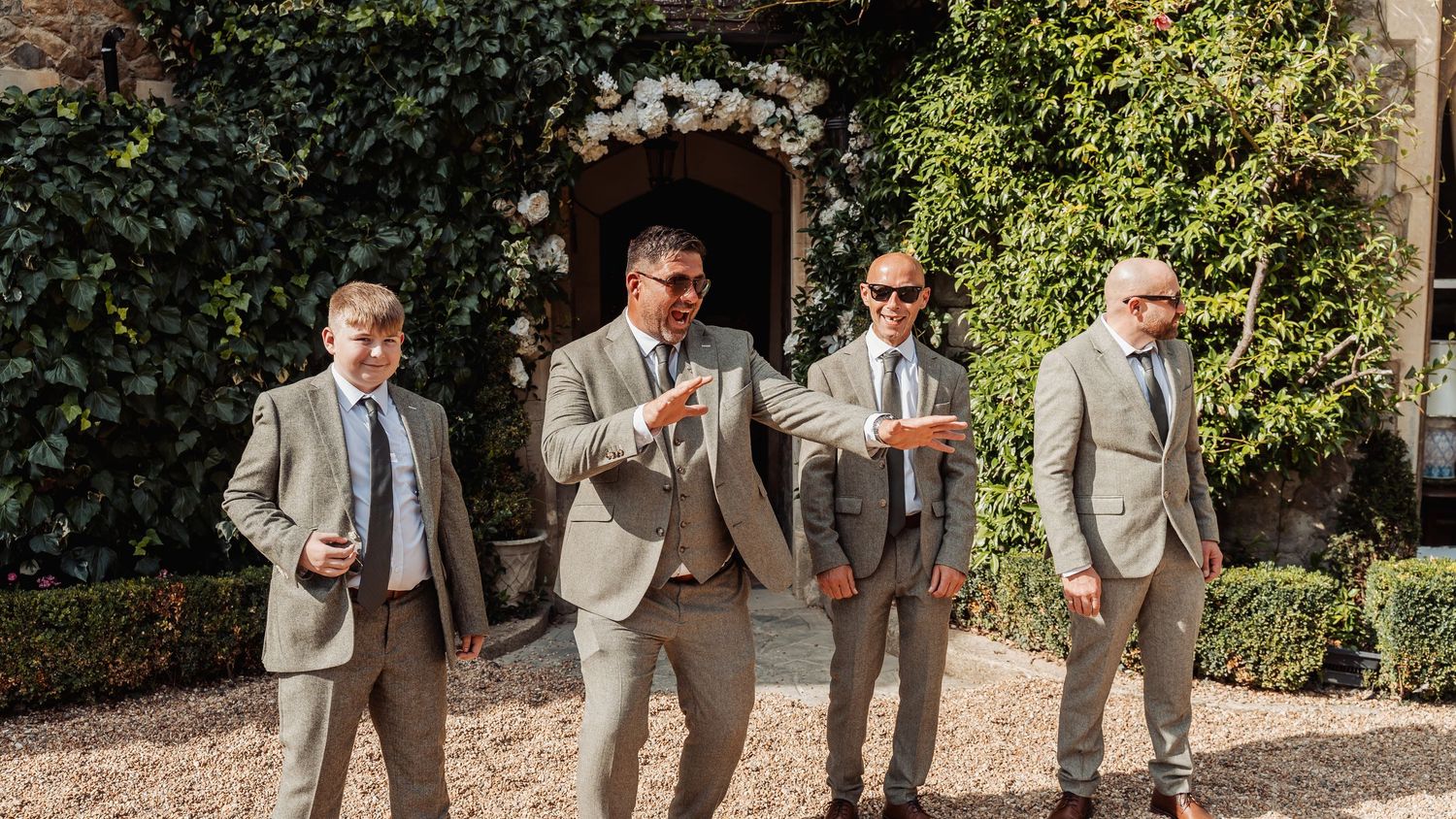 Four groomsmen in matching grey suits posing casually outside ivy-covered stone venue.