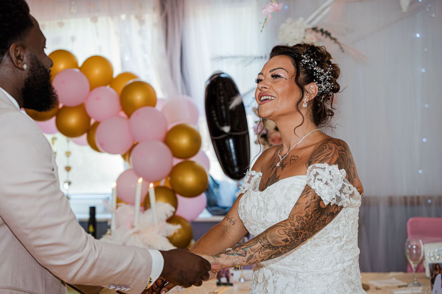Bride with tattoos smiles at groom during wedding reception with pink and gold balloon decorations.