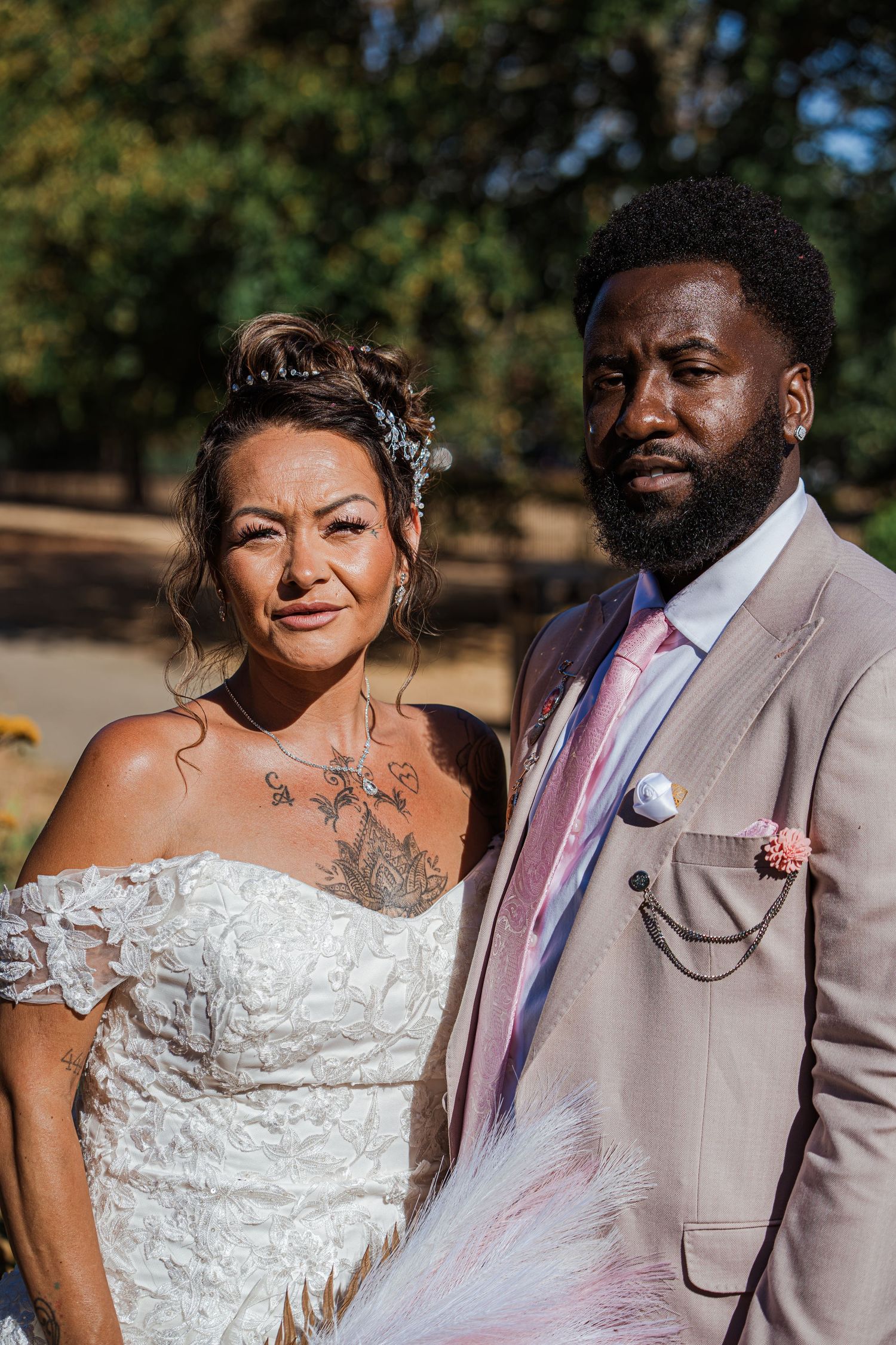 A bride in a white lace dress and groom in a tan suit pose together outdoors at their wedding ceremony.