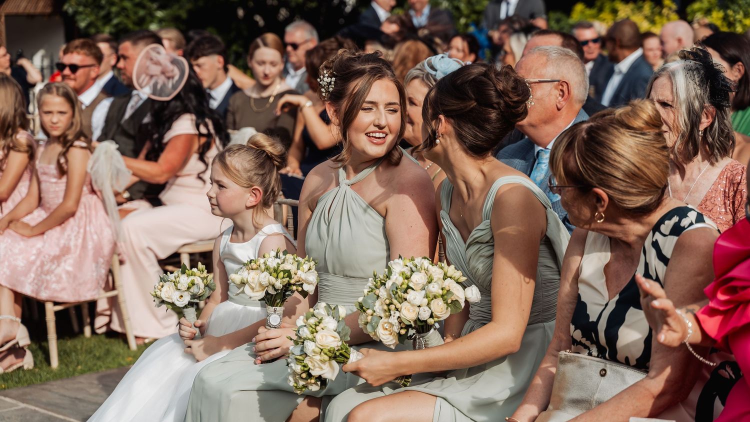 Bridesmaids in sage dresses with white bouquets seated alongside flower girls at ceremony.