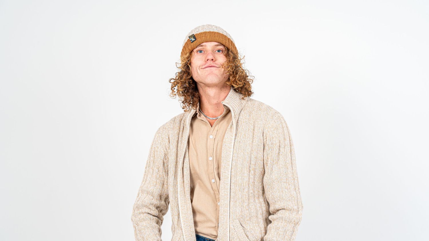 Young man with curly hair wearing a beige knit cardigan and tan beanie, posing against a white background.